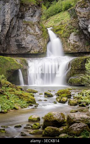 Vista a lunga esposizione di una cascata che si getta in un fiume limpido tra rocce rocciose e scogliere calcaree in una gola verde vicino a Lods, Francia, natura serena Foto Stock