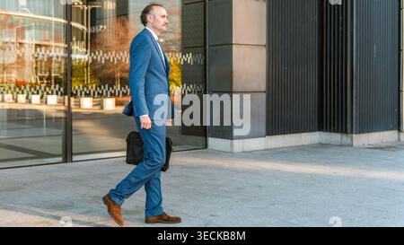 Un uomo d'affari di mezza età con una tuta blu si muove con fiducia verso un moderno edificio di uffici, mostrando un comportamento professionale e determinato su una Foto Stock