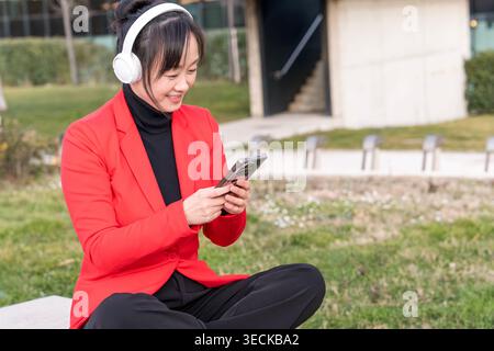 Una donna d'affari di mezza età con un blazer rosso si siede all'aperto, ascoltando musica con le cuffie sorridendo e utilizzando il suo smartphone in una giornata di sole, Foto Stock
