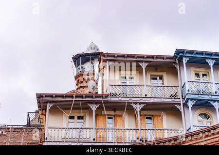 I tradizionali balconi in legno fiancheggiano case colorate nella città vecchia di Tbilisi, Georgia, con una torre in mattoni ornata che si erge dietro sotto un morbido cielo coperto. Foto Stock