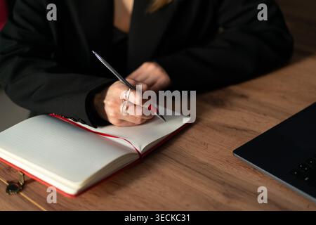 Primo piano di una donna in abito da lavoro che scrive appunti su un notebook aperto su una scrivania di legno accanto a un notebook, catturando pianificazione mirata, produttività, A. Foto Stock