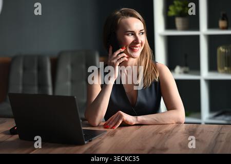 Giovane donna d'affari sorridente che parla con uno smartphone a una scrivania di legno, lavora su un computer portatile in un ufficio moderno. Imprenditrice professionista handlin Foto Stock