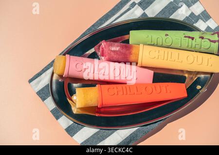 Ghiaccioli di frutta fatti in casa colorati in silicone riutilizzabili su un piatto su un panno a righe, un luminoso spuntino estivo. Foto Stock