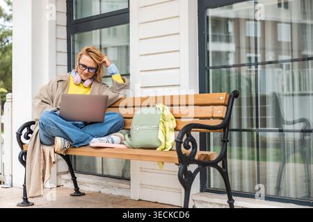 Una donna rilassata siede su una panchina all'aperto, indossa un abbigliamento casual con le cuffie intorno al collo, lavora sul suo laptop, trascorrendo una giornata tranquilla in un Foto Stock