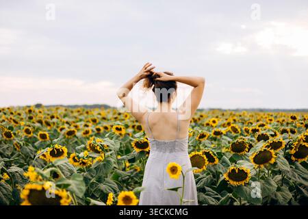 Vista posteriore di una giovane donna con un abito bianco in piedi tra girasoli in fiore, legandosi i capelli e godendosi una tranquilla serata estiva in un vasto paese Foto Stock