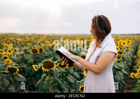 Una donna con un vestito bianco si trova in un campo di girasoli al tramonto, leggendo la Sacra Bibbia. Concetto di fede cristiana pacifica con paesaggio estivo, speranza, Foto Stock
