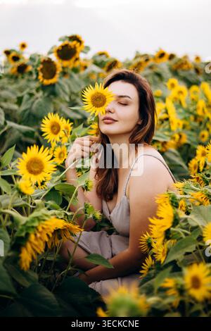 Una giovane donna serena siede tra alti girasoli, tenendo una fioritura vicino al viso con gli occhi chiusi. Calda atmosfera estiva, ritratto naturale, campagna c Foto Stock