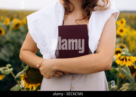 Vista corta di una donna in un campo di girasole che tiene una Sacra Bibbia vicino al suo petto. Scena di campagna estiva che esprime fede cristiana, devozione, p Foto Stock