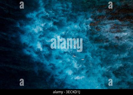 Foto aerea dall'alto verso il basso delle vivide acque turchesi dell'oceano con onde spumeggianti e alcuni surfisti che si spostano vicino a una barriera corallina rocciosa, creando un testo astratto di vista mare Foto Stock