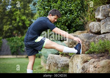 Atleta che si allunga su una roccia in un parco verde, si riscalda per allenarsi e allenarsi. Gli esercizi all'aperto favoriscono flessibilità e mobilità Foto Stock