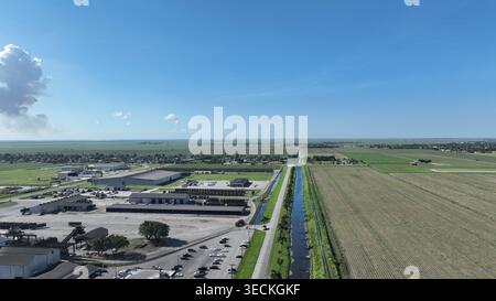 Vista aerea di grandi magazzini industriali e vasti campi agricoli divisi da un canale d'acqua dritto sotto un cielo blu con una grande nuvola bianca. Foto Stock