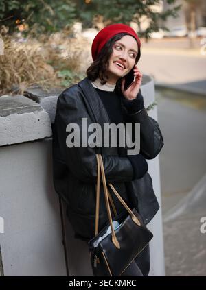 Una giovane donna con capelli scuri che scorre indossa un vivace berretto rosso mentre chatta felicemente sul suo telefono. Si appoggia contro un'elegante cinta muraria della città, trasuda truffa Foto Stock