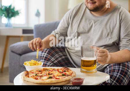 Uomo in sovrappeso che mangia patatine fritte, pizza e birra a casa, seduto sul divano Foto Stock