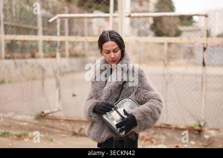 Donna alla moda con un elegante cappotto in pelliccia sintetica che si adatta alla sua borsa in un ambiente urbano circondato da un parco giochi vuoto Foto Stock