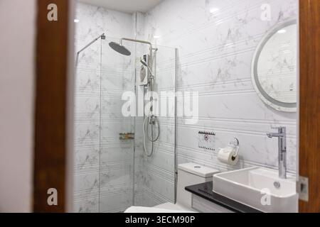 I bagni interni bianchi, luminosi e tranquilli, presentano pareti in piastrelle di marmo, box doccia in vetro, WC pulito, specchio rotondo, lavabo su banco nero Foto Stock