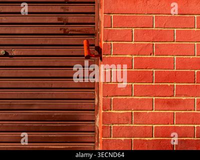 Primo piano di una parete in mattoni dipinti di rosso acceso adiacente a una porta con otturatore in metallo ondulato marrone resistente agli agenti atmosferici. Questo sfondo di texture industriale, acquisito Foto Stock