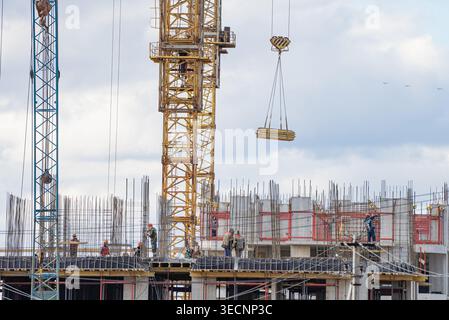 Lavoratori che sviluppano un nuovo e alto edificio in un cantiere affollato, con una grande gru a torre che solleva un fascio di tavole di legno contro un cielo nuvoloso, simboleggiando l'espansione urbana Foto Stock