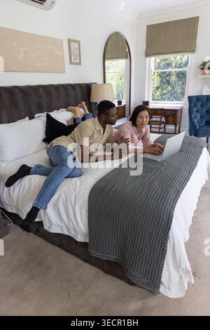 Coppie diverse sdraiate sullo stomaco su un grande letto in una camera da letto luminosa, utilizzando un computer portatile insieme Foto Stock