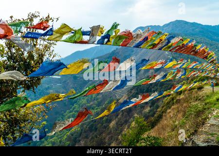 Bandiere di preghiera rettangolari colorate appese in file contro un cielo azzurro pallido, che sventolano nel vento, sagoma di montagna sottostante; viaggio, spiritualità, all'aperto Foto Stock