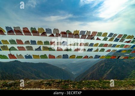 Bandiere di preghiera rettangolari colorate appese in file contro un cielo azzurro pallido, che sventolano nel vento, sagoma di montagna sottostante; viaggio, spiritualità, all'aperto Foto Stock