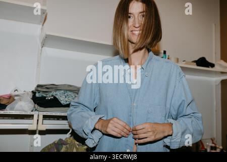 Giovane donna sorridente che abbotta una camicia in denim mentre si veste a casa, in piedi in un guardaroba con scaffali e vestiti, rilassato routi mattutino Foto Stock