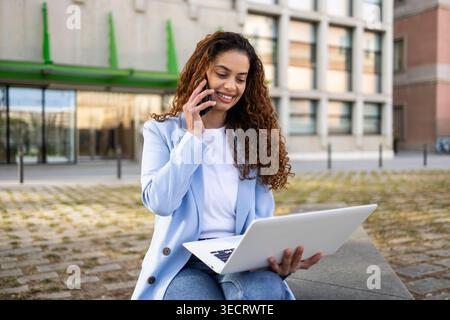 Donna d'affari sorridente in un blazer leggero siede all'aperto vicino a un moderno edificio di uffici, parla su uno smartphone mentre lavora su un laptop e cattura mobi Foto Stock