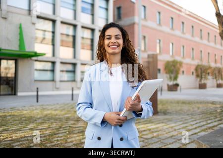 Donna d'affari sorridente in un blazer azzurro che tiene un tablet all'aperto di fronte ai moderni edifici di uffici, conferendo fiducia, leadership e prof Foto Stock