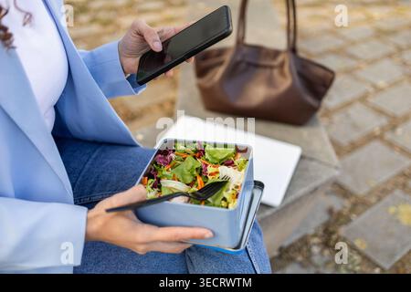 Primo piano di una donna d'affari in un blazer che utilizza uno smartphone mentre mangia un'insalata fresca da un cestino per il pranzo all'aperto, con laptop e borsa nelle vicinanze durante Foto Stock