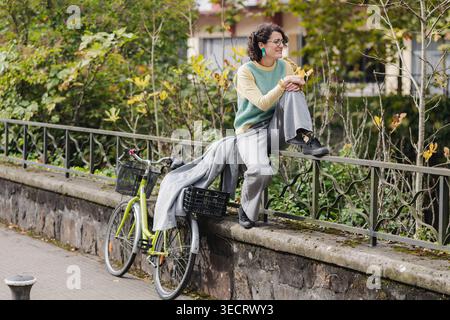 Donna matura con capelli ricci siede su un muro di pietra in un vivace ambiente urbano, godendosi un momento di relax vicino a una bicicletta, abbracciando la vita cittadina e. Foto Stock