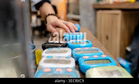 Una fila di timer digitali poggia su un bancone di legno in un ristorante taiwanese, con una mano che si avvicina a uno. I timer sono utilizzati per la precisione nella cottura Foto Stock