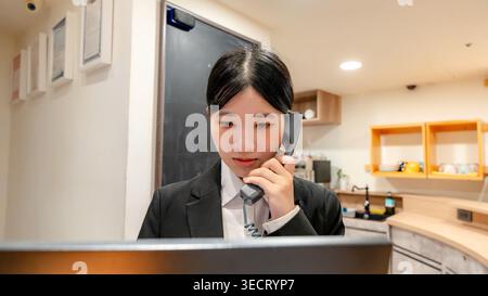 Receptionist taiwanese in una hall moderna che risponde a una telefonata alla reception, focalizzata sul servizio clienti, le prenotazioni e il servizio clienti Foto Stock