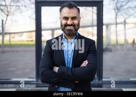 Uomo d'affari arabo fiducioso in una tuta scura sorridente con le braccia incrociate all'aperto, ritratto professionale con sfondo urbano sfocato, leadership, successo Foto Stock