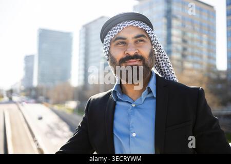 Sicuro uomo d'affari arabo sheikh in keffiyeh tradizionale e abito nero che si posa all'aperto vicino a uno skyline moderno, trasmettendo leadership, successo e urb Foto Stock