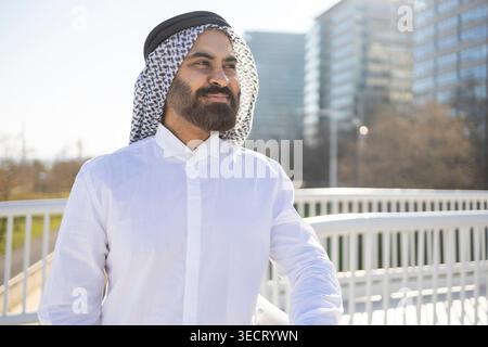 Uomo d'affari arabo sicuro di sé in tradizionale abbigliamento sheikh, con indosso un tobe bianco e un keffiyeh, in piedi all'aperto vicino a una ringhiera con moderni uffici della città dentro Foto Stock