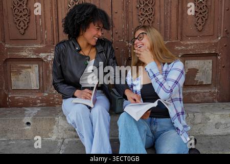Due diverse amiche si siedono su gradini all'aperto, tengono libri, ridono insieme davanti a una vecchia porta di legno, catturano l'amicizia e imparano in modo informale Foto Stock