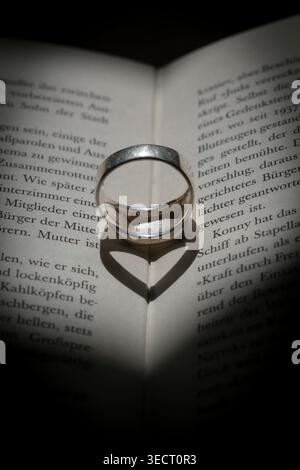Un primo piano di un anello su un libro aperto, la cui ombra forma un cuore perfetto. Foto Stock