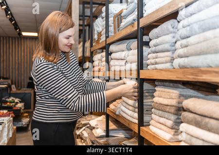 Una donna fa acquisti in un negozio accogliente, esaminando gli asciugamani impilati su scaffali di legno. Indossa una camicia a righe, godendosi la spesa tessile Foto Stock