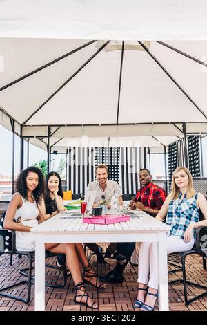 Cinque diversi giovani adulti si siedono insieme a un tavolo sul patio sul tetto sotto un gazebo, sorridendo e chiacchierando sorseggiando un drink, godendosi un'informale riunione estiva Foto Stock