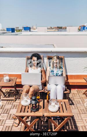 Due giovani donne con gli occhiali da sole siedono su una terrazza soleggiata sul tetto e lavorano su un computer portatile con sedie a sdraio. Ambiente di lavoro remoto all'aperto con luce estiva, bevande, Foto Stock