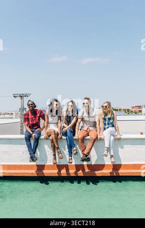 Cinque giovani adulti diversi siedono su una sporgenza sul tetto, sorridendo e chiacchierando in una giornata di sole. Moda informale, cielo blu, sfondo urbano e weekend rilassato Foto Stock
