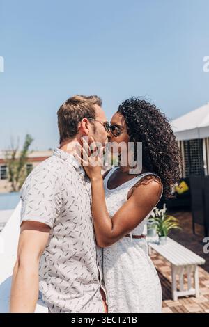 Coppia romantica che si baciano all'aperto in una giornata estiva di sole, indossando occhiali da sole in abiti casual, celebrando l'amore e l'intimità su una luminosa terrazza con c Foto Stock