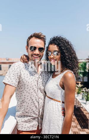 Coppia felice e variegata che posa con gli occhiali da sole su una terrazza soleggiata sul tetto, godendosi un'atmosfera estiva, lo skyline della città sullo sfondo, rilassata vita di viaggio Foto Stock