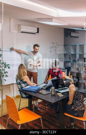 Diversi colleghi si riuniscono in una moderna sala riunioni dell'ufficio mentre il relatore spiega un piano su una lavagna a fogli mobili, mentre i colleghi collaborano con computer portatili e. Foto Stock