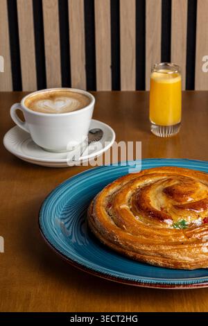 Accogliente colazione mattutina su un tavolo da caffe' con un latte in una tazza bianca, un bicchiere di succo d'arancia e una pasticceria a spirale smaltata su un piatto blu, caldo e. Foto Stock