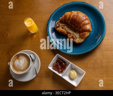 Vista dall'alto di una semplice colazione mattutina su un tavolo di legno con croissant su un piatto blu, cappuccino con latte art, succo d'arancia, burro e marmellata. Foto Stock