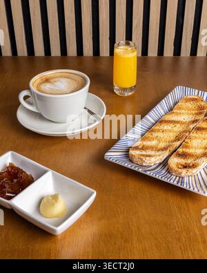 Colazione al tavolo con cappuccino in una tazza bianca, pane tostato su un piatto a strisce, burro e marmellata e un piccolo bicchiere di succo d'arancia su un legnoso Foto Stock