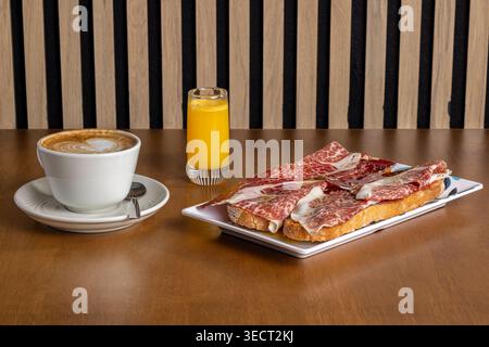 Colazione mattutina su un tavolo di legno con caffè e succo d'arancia, con pane tostato con prosciutto spagnolo su un piatto bianco, ideale per caffè o. Foto Stock