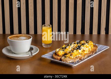 La colazione mattutina, servita su un tavolo, comprende una tazza di latte, un bicchiere di succo d'arancia e pane tostato con uova strapazzate e semi Foto Stock