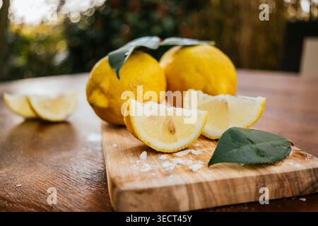 Primo piano di limoni freschi con foglie verdi e cunei affettati su un tagliere di legno, luce naturale e morbido sfondo bokeh, perfetto per il cibo, Foto Stock