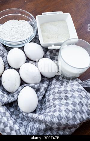 Deporre gli ingredienti da forno su un panno a scacchi, uova fresche, un recipiente di farina, zucchero bianco in un piatto e un bicchiere di latte su un tavolo di legno. Foto Stock